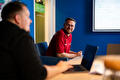 Een man met een rode polo zit aan een tafel met een open laptop en kijkt naar een gesprekspartner op de voorgrond. Op de achtergrond hangt een whiteboard met onduidelijke aantekeningen.