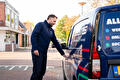 Een man in donkere kleding staat naast een blauwe bestelbus en raakt met zijn hand de zijkant aan. Op de bus staan gedeeltelijk zichtbare woorden en gekleurde stippen.